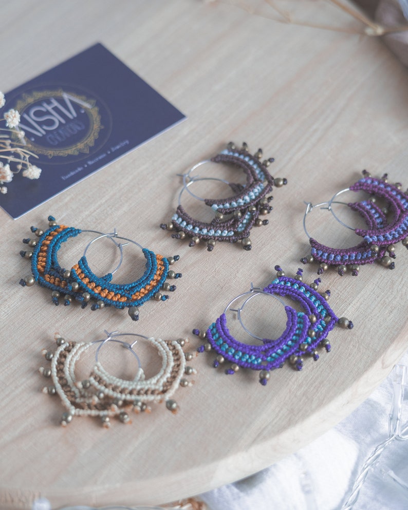 May include: Five pairs of handmade macrame hoop earrings with different colored threads and brass beads. The earrings are displayed on a wooden surface.