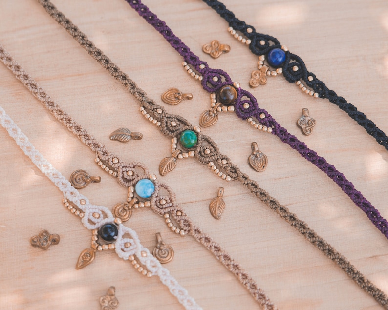 May include: Four macrame anklets with different colored cords and gemstone beads. The anklets are made with a variety of beads, including turquoise, lapis lazuli, tiger's eye, and onyx. Each anklet has a small brass charm hanging from it.