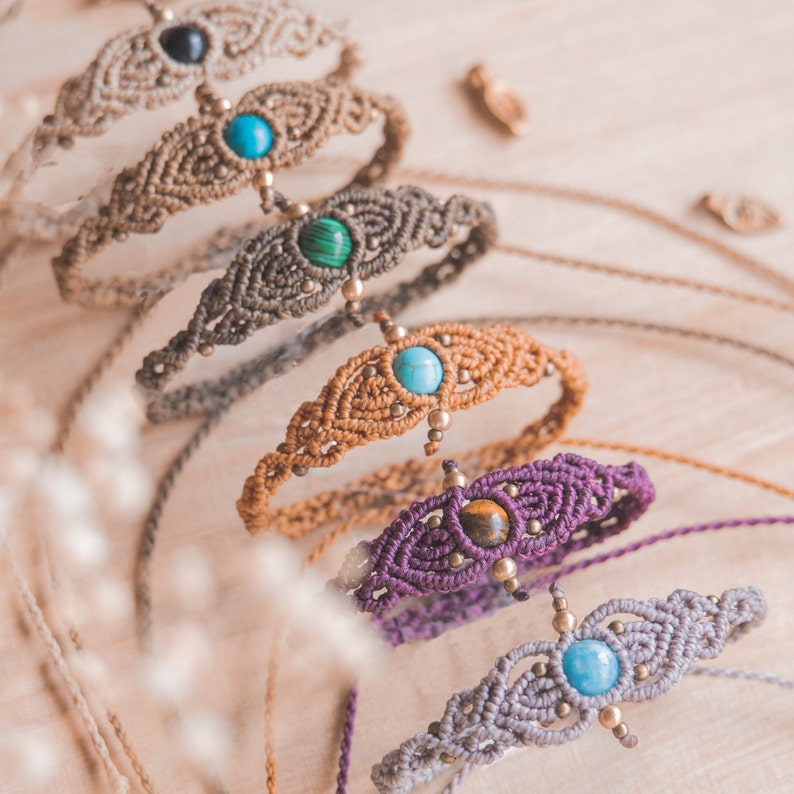 May include: A set of five macrame bracelets with different colored stones. Each bracelet features a unique stone in the center, including turquoise, malachite, tiger's eye, and onyx. The bracelets are made with brown, purple, gray, and beige cords.