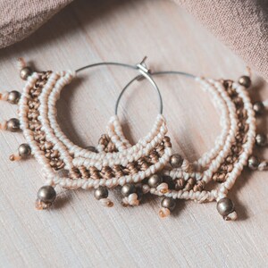 May include: A pair of large hoop earrings made with brown and white macrame cord and gold beads. The earrings are hanging on a light wood surface.