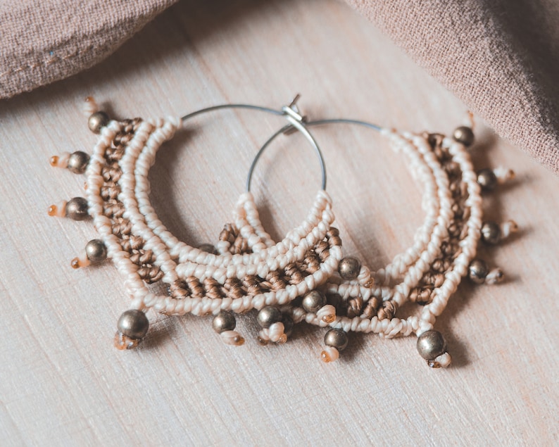 May include: A pair of large hoop earrings made with brown and white macrame cord and gold beads. The earrings are hanging on a light wood surface.