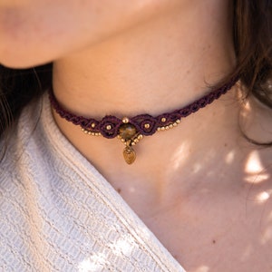 May include: A burgundy macrame choker necklace with a gold pendant and a tiger's eye stone.