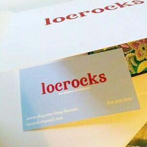 May include: A blue business card with the text "locrocks an evessa enterprise" in red. The card also includes the website address "www.etsy.com/shop/locrocks" and the email address "locrocks@gmail.com". The phone number "202.256.5236" is printed in white on the card.
