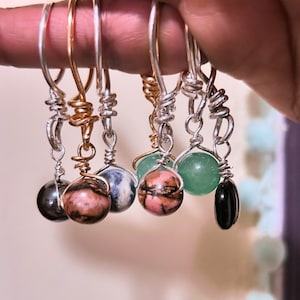May include: A collection of handmade earrings with various gemstone beads. The earrings feature silver and gold-colored wire, with beads in shades of black, pink, blue, green, and a black teardrop shape. The earrings are held up by a finger.