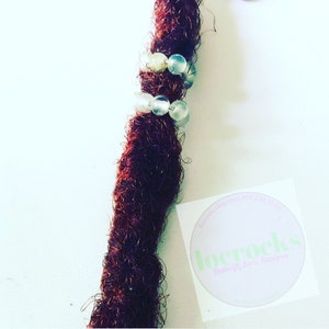 May include: Close-up of a single brown dreadlock with two rows of clear beads. The dreadlock is tied with a clear elastic band. A sticker with the text "locrocks Rubberfly Locs Designer" is visible in the background.