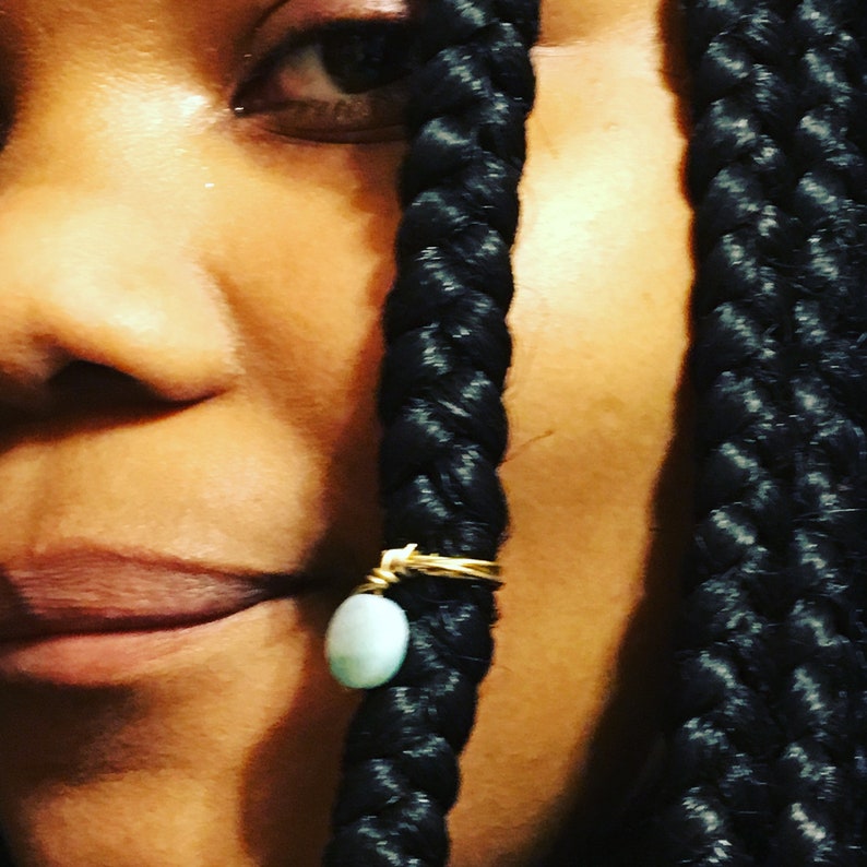 May include: Close-up of a person's face with black braided hair. A gold ring with a light blue oval bead is in the braid.