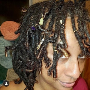K&ouml;nnte beinhalten: Nahaufnahme von einer Person mit Dreadlocks, die mit goldenem und perlenbesetztem Haarschmuck verziert sind. Die Dreadlocks sind in einer lockigen Form gestylt und der Haarschmuck ist im gesamten Haar verteilt.