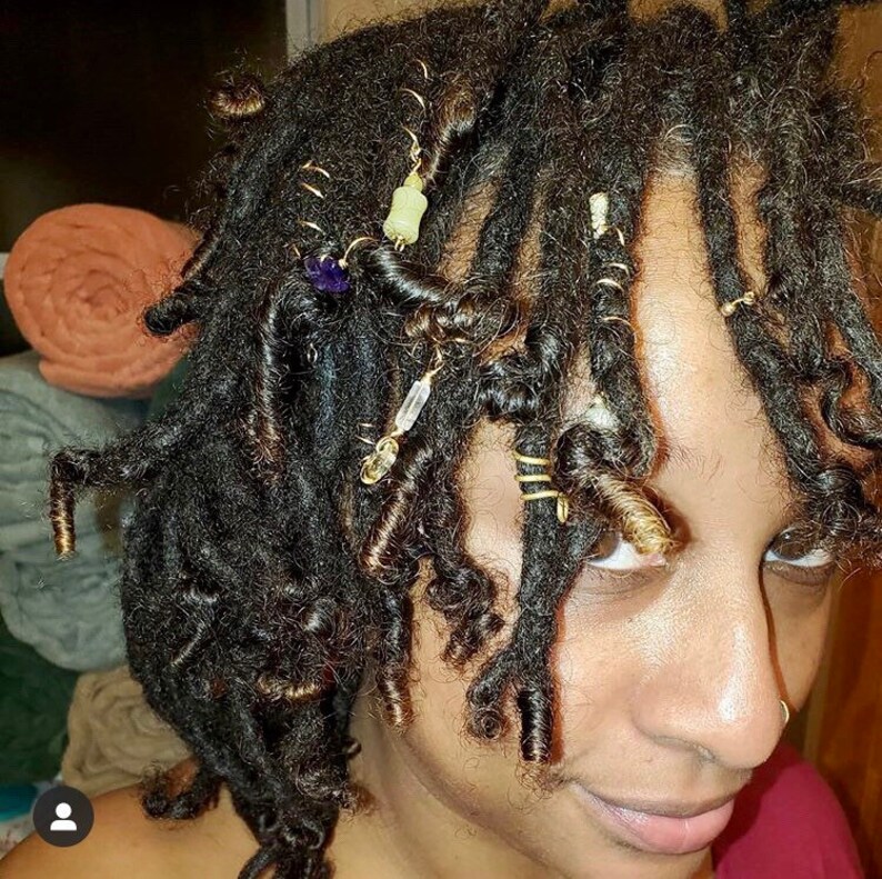 K&ouml;nnte beinhalten: Nahaufnahme von einer Person mit Dreadlocks, die mit goldenem und perlenbesetztem Haarschmuck verziert sind. Die Dreadlocks sind in einer lockigen Form gestylt und der Haarschmuck ist im gesamten Haar verteilt.