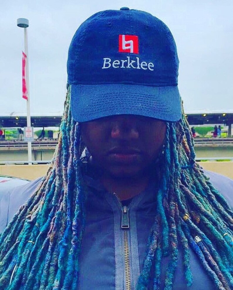 K&ouml;nnte beinhalten: Eine Person mit langen blauen und gr&uuml;nen Dreadlocks tr&auml;gt eine dunkelblaue Baseballkappe mit einem wei&szlig;en Logo, auf dem "Berklee" steht, und einem roten Quadrat mit dem Buchstaben "B" darin.