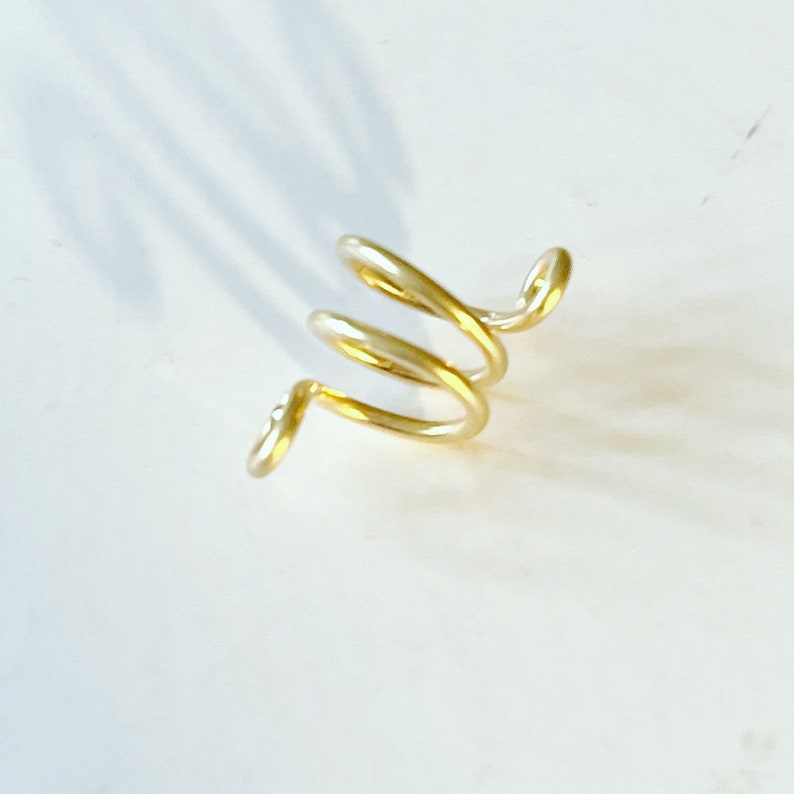 May include: A gold-colored spiral ear cuff made of wire. The cuff is designed to wrap around the ear and has a simple, modern look.