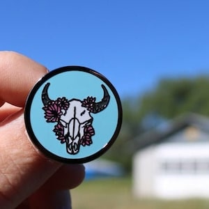 Puede incluir: Un pin de esmalte azul con un diseño de cráneo de toro blanco. El cráneo está adornado con flores rosas.
