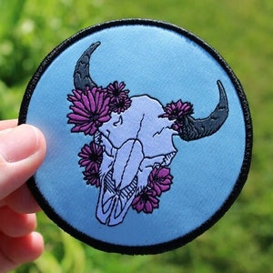 May include: A blue embroidered patch with a white skull and black horns surrounded by purple flowers.