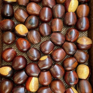 May include: A close-up of a basket filled with many small, round, wooden objects. The objects are a rich reddish-brown color with darker brown streaks. Some of the objects have a lighter brown band around the middle.