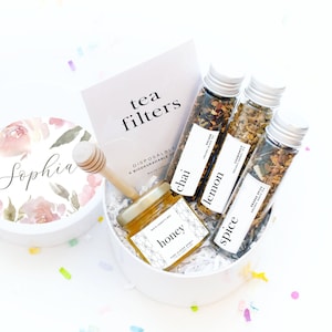May include: A white gift box with a floral design and the name "Sophia" on the lid. The box contains a small jar of honey, four glass vials of tea, and a wooden honey dipper. The tea flavors are chai, lemon, spice, gold, frank, and myrrh. The box is labeled "White Confetti Box".