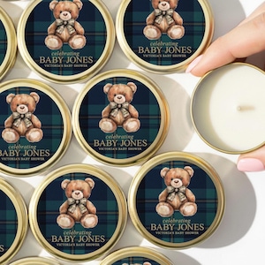 May include: Small, round candles in gold-colored tins. Each tin features a teddy bear illustration on a navy blue and green plaid background, with the text "celebrating BABY JONES". A hand holds one candle.