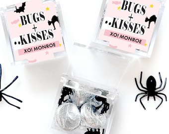 May include: Three clear square plastic favor boxes filled with silver foil-wrapped candies. Each box features a light pink label with black text reading BUGS + KISSES and XO! MONROE, decorated with black bat and cat silhouettes and yellow stars.