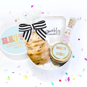 May include: A white gift box with a blue lid that says "Happy Birthday Jenna". The box contains cookies, a small candle that says "Make a Wish", and a small bottle with a pink label that says "Dream Fairy Dust".