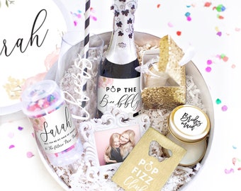 May include: A white gift box filled with party favors for a bridal shower. The box includes a mini bottle of champagne with a label that says "Pop the Bubbly", a gold bottle opener with the text "Pop Fizz Clink", a small gold container with the text "Bridesmaid", a confetti popper with the text "Sarah", a small gold frame with the text "Bride Tribe", a gold fortune cookie, a gold glitter box, and a clear glass with the text "Cheers".