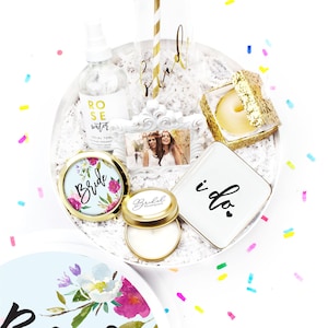 May include: A white gift box filled with bridal shower gifts, including a glass of champagne, a compact mirror, a candle, a small bottle of rose water, a tissue box, and a small white plate with the words "i do" on it. The box is surrounded by confetti.