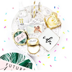 May include: A collection of bridal-themed gifts, including a champagne flute with "bride" in gold script, a small dish with "i do", and a compact mirror with "bride" on it. The items are arranged on a white surface.