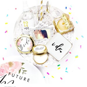 May include: A white gift box filled with bridal shower gifts, including a compact mirror, a candle, a glass of champagne, a picture frame, a tissue box, a bottle of rose water, and a small plate with the words "i do" on it. The box is surrounded by colorful confetti.