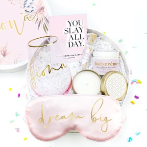 K&ouml;nnte beinhalten: Eine rosa und wei&szlig;e Geschenkbox mit verschiedenen Artikeln, darunter ein Glas mit goldfarbenem Rand, eine Kerze, eine Schlafmaske und ein Tiegel mit K&ouml;rpercreme. Die Box ist mit dem Namen "Fiona" und dem Spruch "Dream Big" verziert. Die Box enth&auml;lt au&szlig;erdem sechs Affirmationskarten mit dem Text "You Slay All Day."
