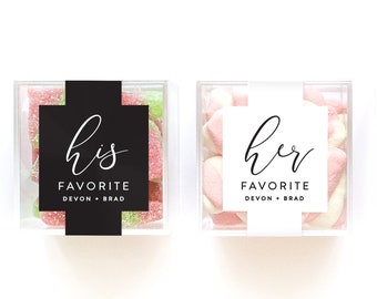 May include: Two clear square boxes filled with candy. The box on the left has green and pink candy and a black label that says "his favorite Devon + Brad". The box on the right has white and pink candy and a white label that says "her favorite Devon + Brad".