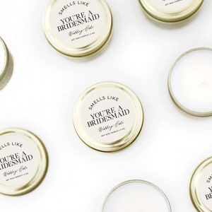 May include: A set of eight small gold candle jars with white lids. Each jar has a black and white label that reads "Smells Like You're a Bridesmaid Wedding Cake".