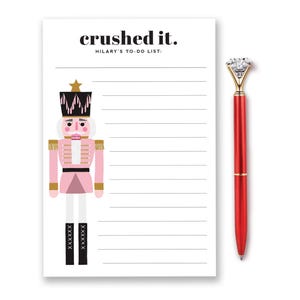 May include: A white notepad with the text "crushed it." and a pink nutcracker illustration. A red pen with a diamond-shaped top is next to the notepad. The notepad is a to-do list.