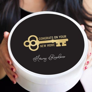 May include: A white round gift box with a black lid. The lid has a gold key design and the text "Congrats on your new home Harvey Residence".