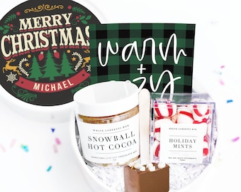 May include: A white gift box filled with hot cocoa mix, a chocolate spoon, holiday mints, and a card that says "warm + cozy". The lid says "Merry Christmas" with a festive design. Confetti is scattered around the box.