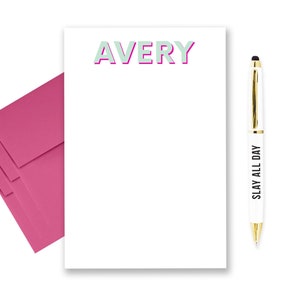 May include: A white notepad with the name "AVERY" printed in light green with a pink outline. A pink envelope is stacked on the left side of the notepad. A white pen with a gold top and the text "SLAY ALL DAY" printed on the side is on the right side of the notepad.