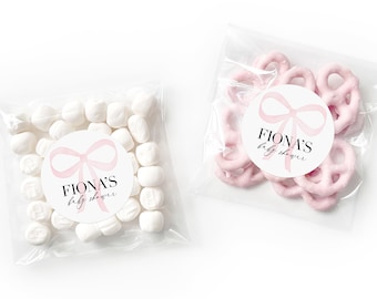 May include: Two clear cellophane bags filled with party favors. The bag on the left contains white candy. The bag on the right contains pink pretzel bites. Both bags have a round sticker with a pink bow and the text "Fiona's Baby Shower".