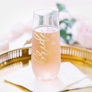 May include: A clear glass champagne flute with a gold inscription that reads "bride" filled with a pink beverage. The flute is sitting on a gold tray with a white napkin.