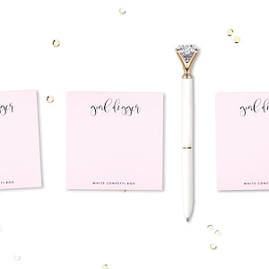 May include: Three pink notepads with the text "goal digger" written in black cursive on each notepad. The notepads are on a white surface with gold confetti scattered around them. A white pen with a gold diamond-shaped top is in the center of the image.