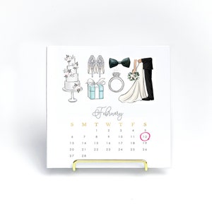 May include: A square calendar with a gold stand features wedding-themed illustrations, including a cake, shoes, a bow tie, a ring, and a bride and groom. The month of February is displayed, with the 12th circled in pink.