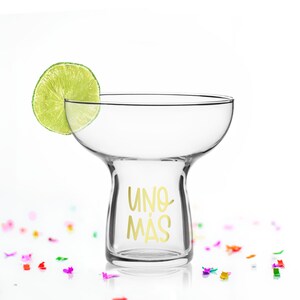 May include: A clear glass margarita glass with a gold inscription that reads "UNO MAS". A lime wedge sits on the rim of the glass. The glass is surrounded by colourful confetti.