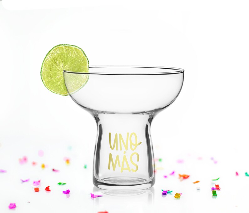 May include: A clear glass margarita glass with a gold inscription that reads "UNO MAS". A lime wedge sits on the rim of the glass. The glass is surrounded by colourful confetti.