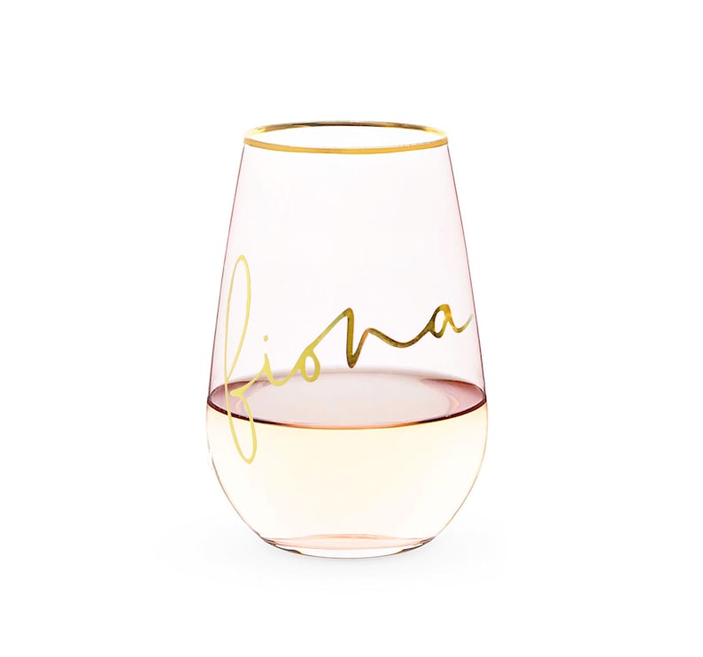 K&ouml;nnte beinhalten: Ein klares Weinglas aus Glas mit goldenem Rand und dem Namen "Fiona" in goldener Schrift auf der Seite. Das Glas ist mit einem hellen Getr&auml;nk gef&uuml;llt.