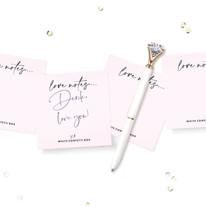 May include: Four pink note cards with the text "love notes..." printed on them. One card has the additional text "Derik, love you! xo". A white pen with a gold-toned diamond-shaped top is lying on the cards.