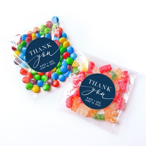Peut inclure: Deux sacs en plastique transparents remplis de bonbons. Le sac &agrave; gauche contient des M&Ms color&eacute;s. Le sac &agrave; droite contient des bonbons g&eacute;lifi&eacute;s color&eacute;s. Les deux sacs ont des autocollants ronds bleus avec du texte blanc qui dit "THANK you" avec les noms "KATE + LEO" et la date "JUNE 18, 2025".