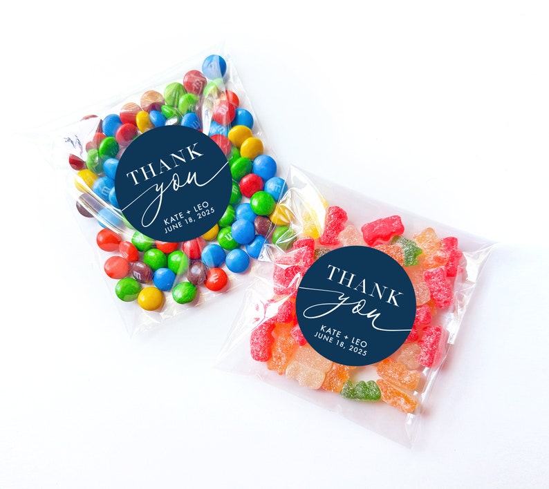 Peut inclure: Deux sacs en plastique transparents remplis de bonbons. Le sac &agrave; gauche contient des M&Ms color&eacute;s. Le sac &agrave; droite contient des bonbons g&eacute;lifi&eacute;s color&eacute;s. Les deux sacs ont des autocollants ronds bleus avec du texte blanc qui dit "THANK you" avec les noms "KATE + LEO" et la date "JUNE 18, 2025".