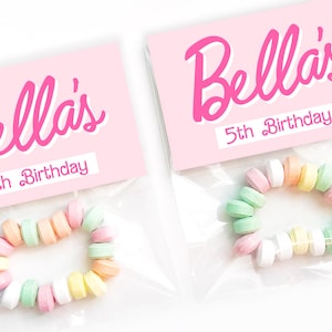 May include: Two clear plastic bags each contain a candy bracelet and a pink label. The labels read "Bella's 5th Birthday" in pink script. The bracelets are made of pastel-colored candies.