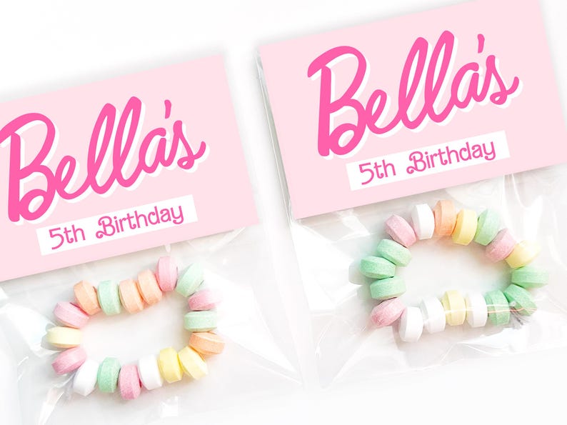 May include: Two clear plastic bags each contain a candy bracelet and a pink label. The labels read "Bella's 5th Birthday" in pink script. The bracelets are made of pastel-colored candies.