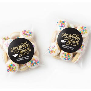 May include: Two clear cellophane bags filled with cookies. Each cookie is round with white icing and colorful sprinkles. A black circular sticker on each bag reads "Congrats Grad" with a graduation cap graphic and the name "Candace James CLASS OF 2026."