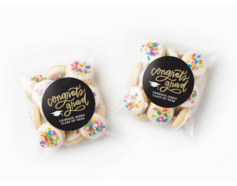 May include: Two clear cellophane bags filled with cookies. Each cookie is round with white icing and colorful sprinkles. A black circular sticker on each bag reads "Congrats Grad" with a graduation cap graphic and the name "Candace James CLASS OF 2026."