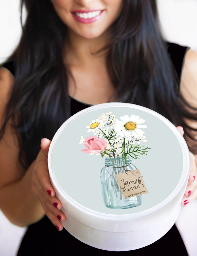 Puede incluir: Una caja de regalo redonda blanca con una tapa azul claro que presenta un tarro de vidrio lleno de margaritas blancas, rosas rosadas y vegetaci&oacute;n. El tarro tiene una etiqueta marr&oacute;n que dice "James Residence Happy New Home".