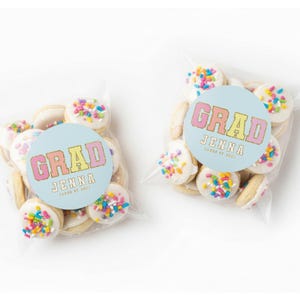 May include: Two clear cellophane bags filled with cookies. Each cookie is round, topped with white frosting and colorful sprinkles. A light blue circular sticker on each bag reads "GRAD JENNA CLASS OF 2023" in pink, yellow, and orange.