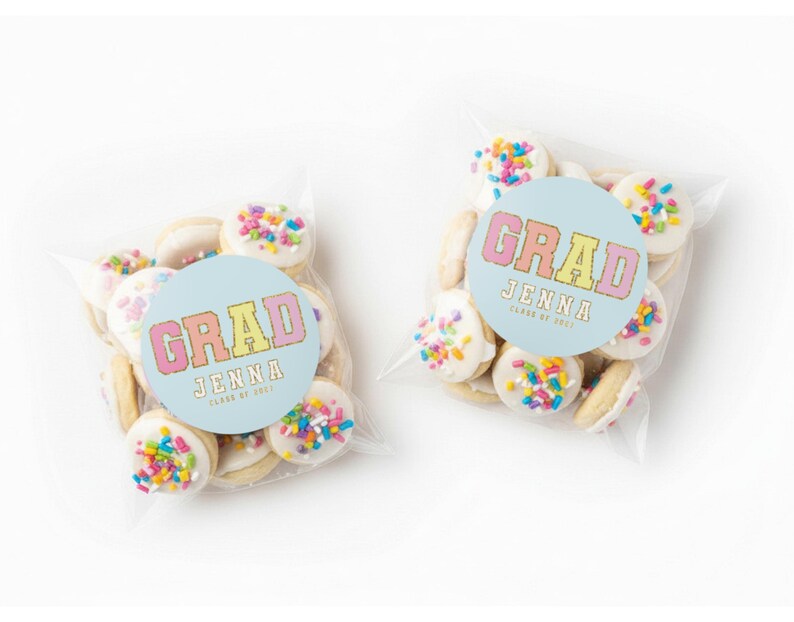 May include: Two clear cellophane bags filled with cookies. Each cookie is round, topped with white frosting and colorful sprinkles. A light blue circular sticker on each bag reads "GRAD JENNA CLASS OF 2023" in pink, yellow, and orange.
