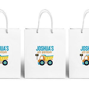 May include: Three white paper gift bags with blue and yellow construction truck themed labels. The labels say "Joshua's 8th Birthday".